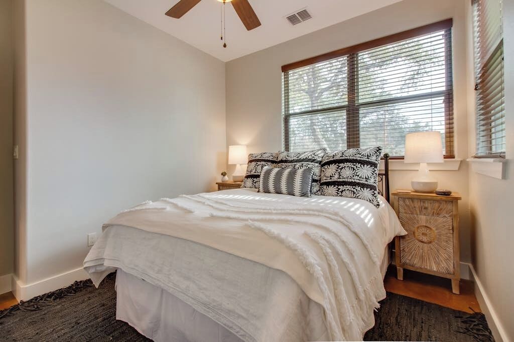 A bedroom with a bed, two bedside tables, a window with blinds, a ceiling fan, and a nightstand with a lamp.