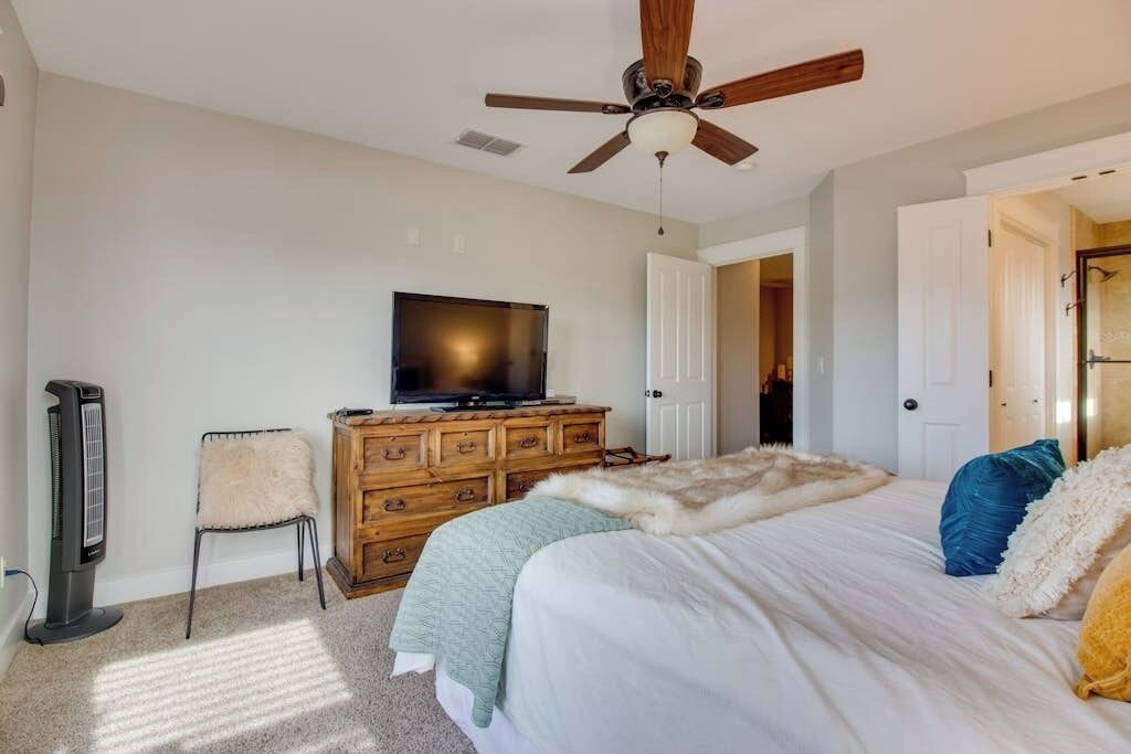A bedroom with a bed, a wooden dresser, a television, a ceiling fan, and a door leading to another room.