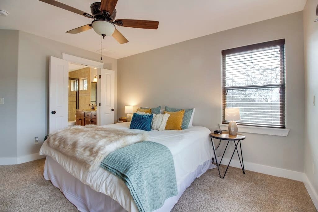 Un dormitorio con cama, ventilador de techo, ventana con persianas y mesita de noche con lámpara.