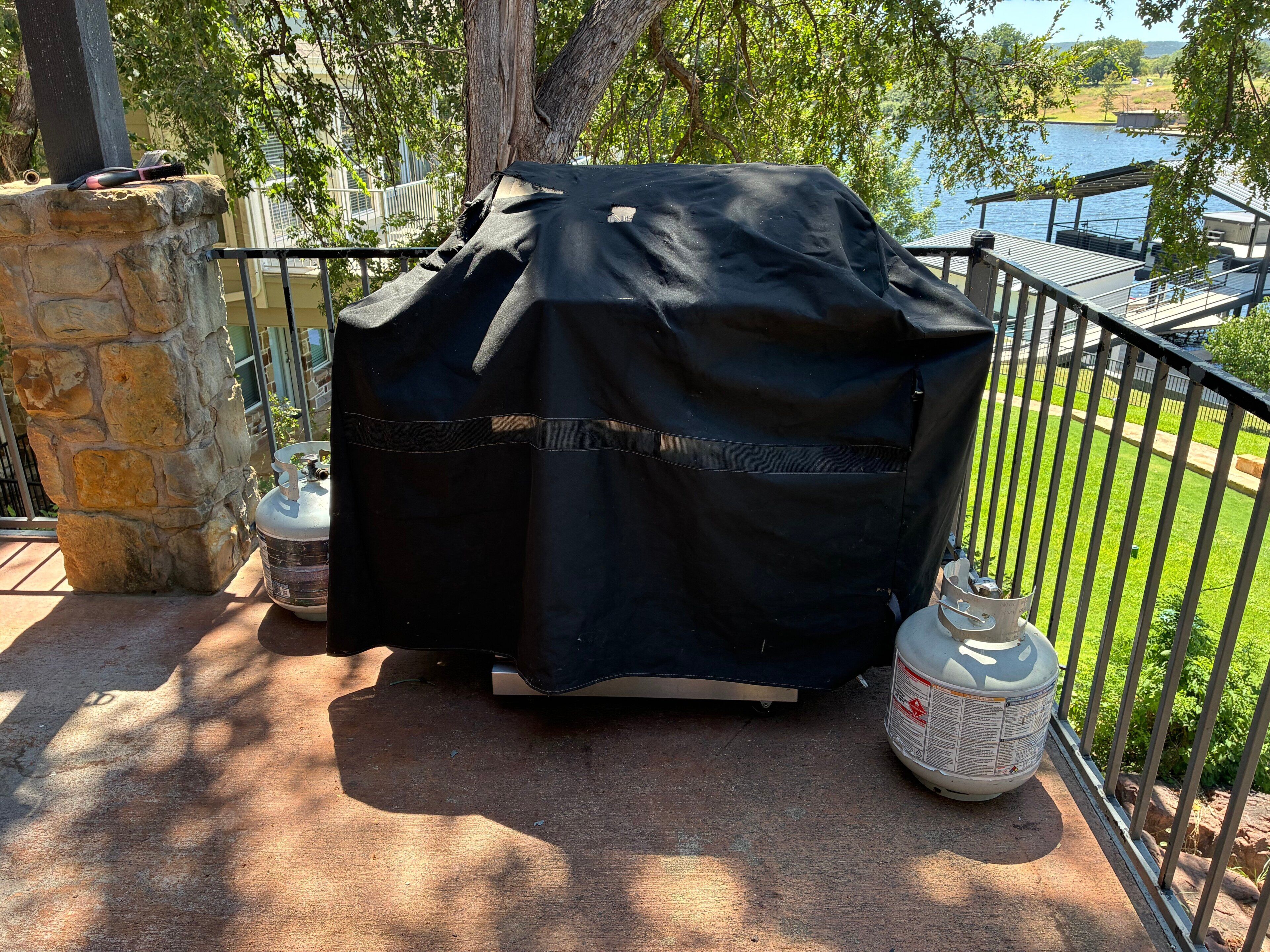 A barbecue grill covered with a black cloth, a propane tank, and a stone pillar.