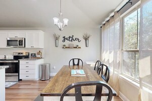 Uma sala de jantar com uma mesa de madeira, cadeiras, um lustre e uma cozinha ao fundo.