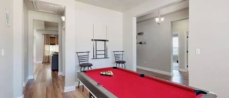 A pool table in a room with wooden flooring, a ceiling fan, and a hallway leading to another room.