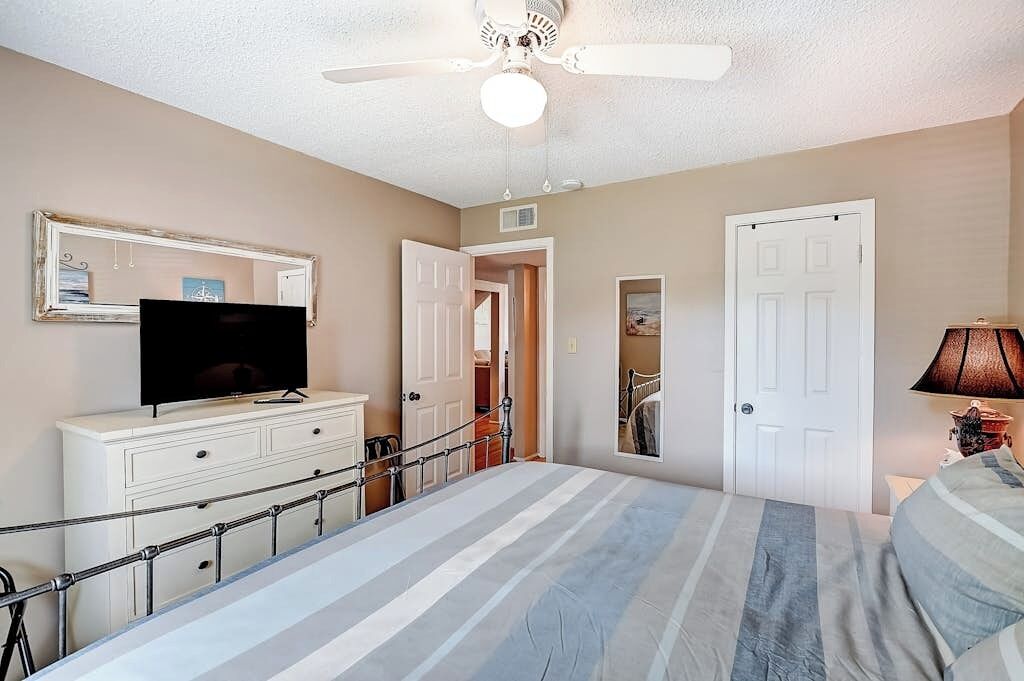 A bedroom with a bed, a television on a dresser, a ceiling fan, and a door leading to another room.