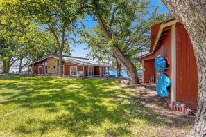A rustic cabin with a stone foundation, a red exterior, and a large tree in the foreground. - Multiple, Dog-friendly Waterfront Homes With Lake LBJ Views - Snowbird-friendly (Kingsland)
