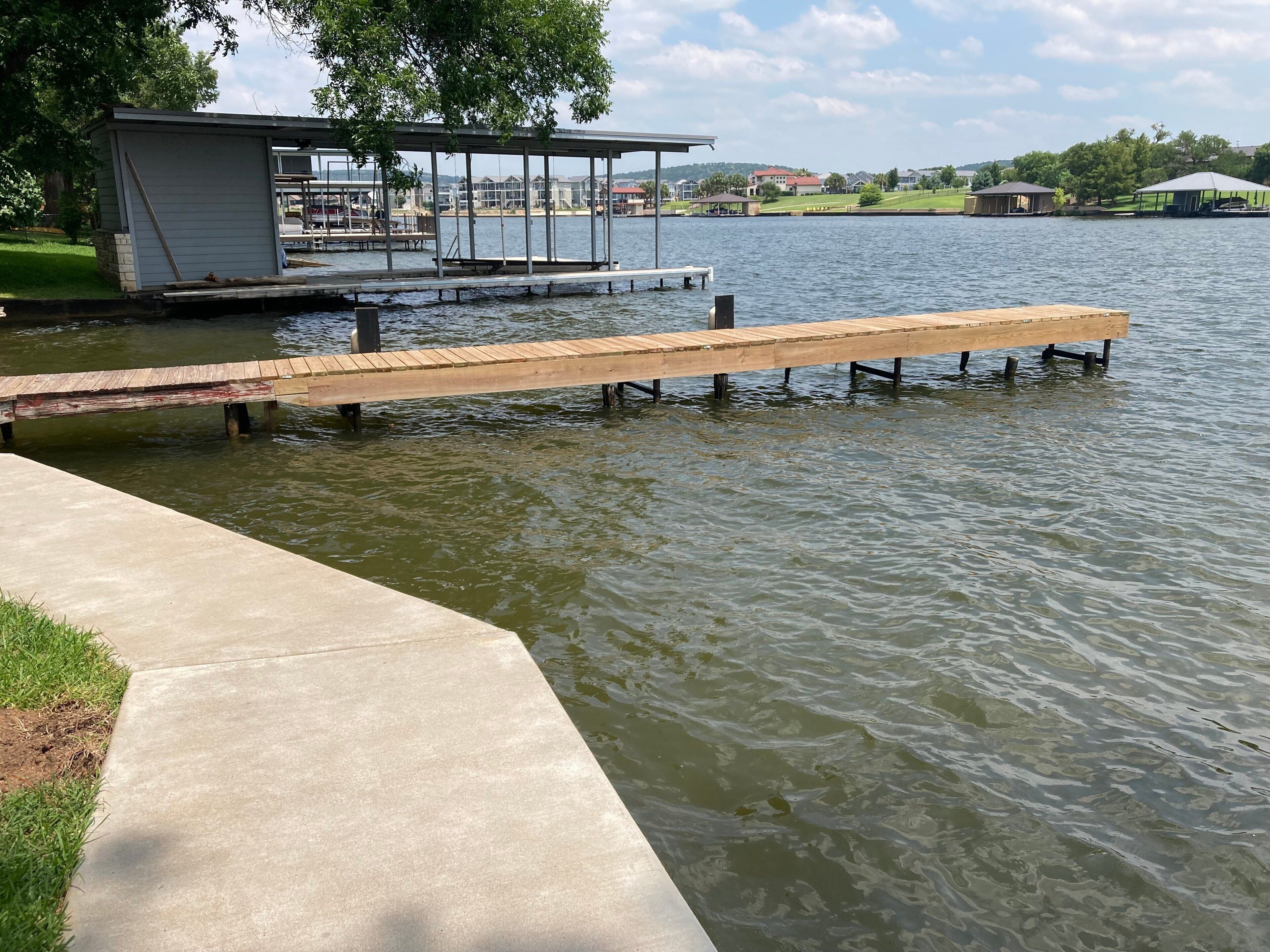 A concrete dock extending into a lake, a small pavilion, and a grassy area with a building in the background.