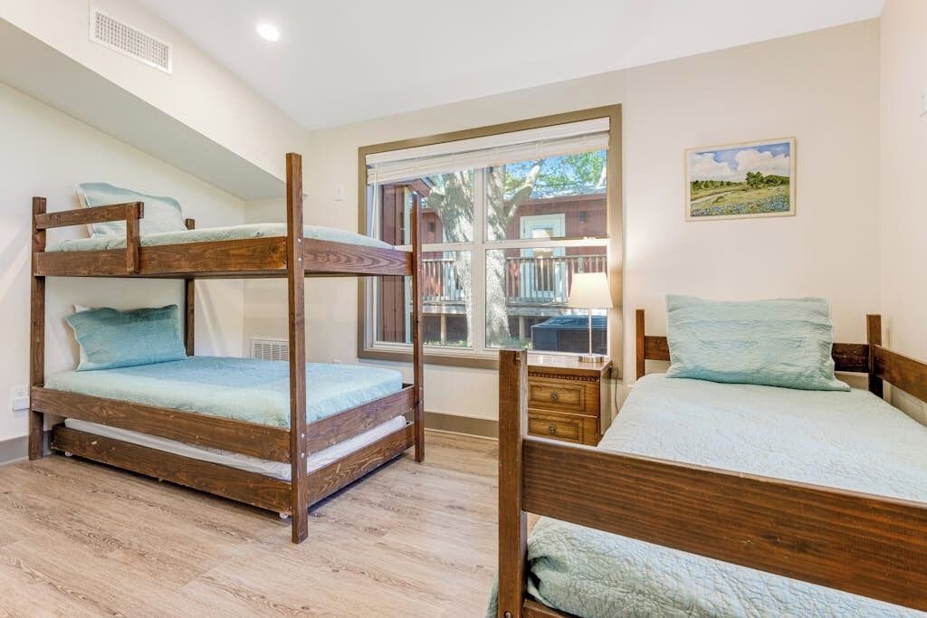 A room with a bunk bed, a single bed, a window with a view, and a small table with a lamp.