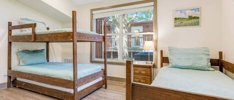 A room with a bunk bed, a single bed, a window with a view, and a small table with a lamp.