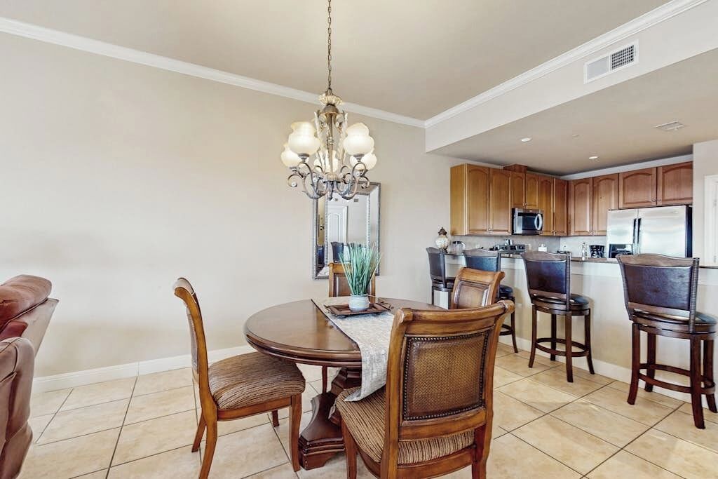 A dining area with a chandelier, wooden chairs, a round table, and a kitchen in the background.