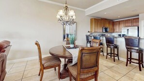 A dining area with a chandelier, wooden chairs, a round table, and a kitchen in the background. - Waterfront Condo With Furnished Balcony, Central AC, & Washer/dryer (Horseshoe Bay)