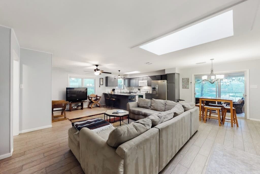 Una sala moderna con un sofá seccional, un comedor con mesa y sillas, una cocina con refrigerador y microondas, y un tragaluz grande.