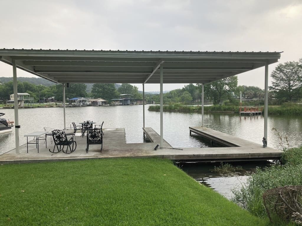 Un muelle cubierto con sillas y una mesa, un césped y un lago tranquilo con barcos al fondo.