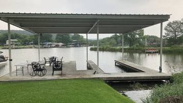 A covered dock with chairs and a table, a grassy area, and a calm lake with boats in the background.