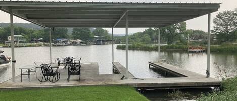 Un muelle cubierto con sillas y una mesa, un césped y un lago tranquilo con barcos al fondo.