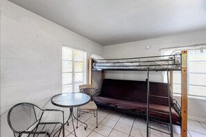 Ein Etagenbett mit Matratze, ein runder Tisch mit Stühlen und ein Sofa in einem Raum mit schlichten Wänden und Fenstern.