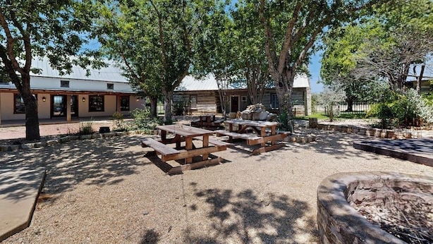 Un área de patio al aire libre con mesas de picnic, un hogar de piedra y un edificio con techo blanco al fondo.