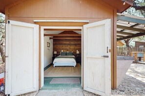 Uma cabana de madeira com a porta aberta, revelando um quarto com cama, almofadas e uma mesinha de cabeceira.