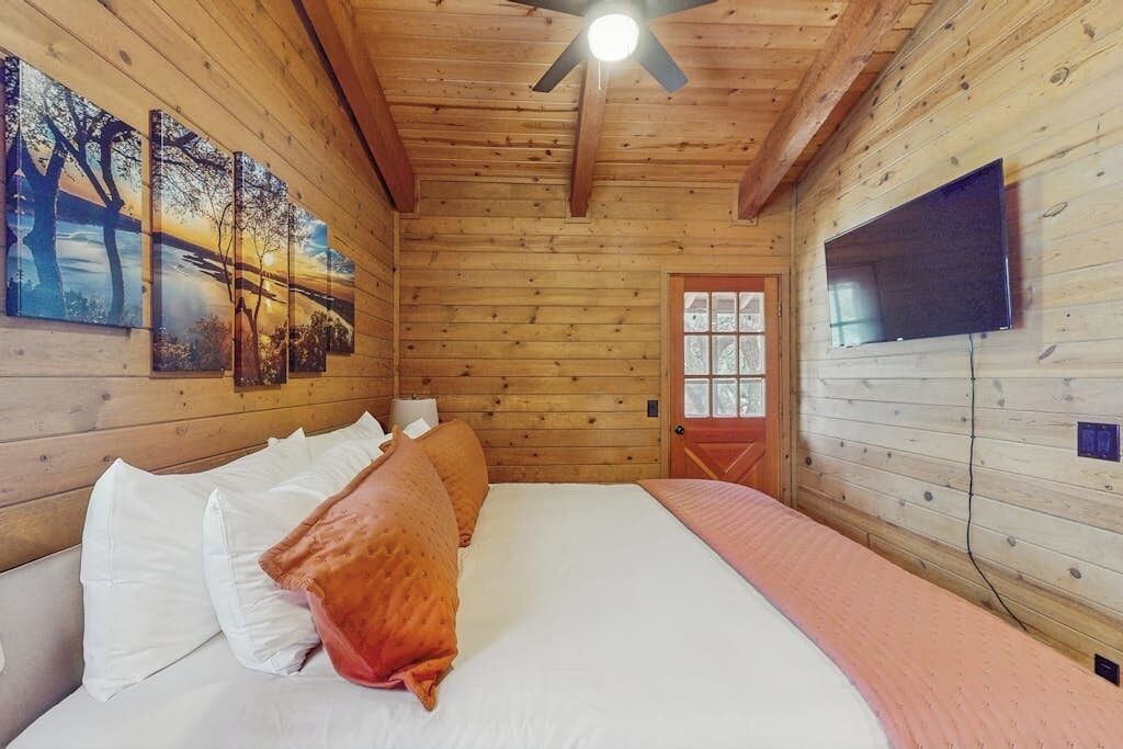 A wooden cabin room with a bed, a ceiling fan, a TV mounted on the wall, and a window with a view.