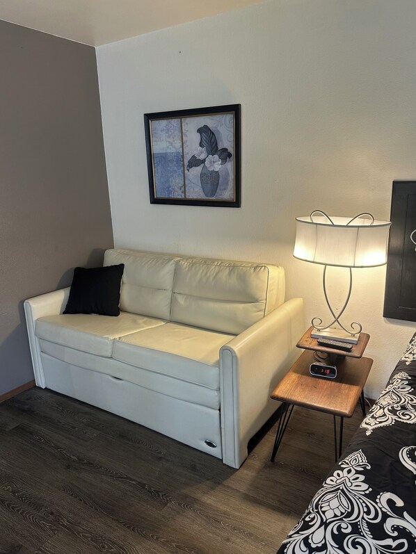 Una habitación con un sofá blanco, una mesita de noche de madera con una lámpara, una obra de arte floral enmarcada en la pared y una cama con colcha estampada.