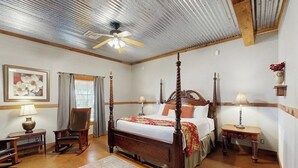 A four-poster bed with a wooden headboard, a ceiling fan, a framed floral artwork, a rocking chair, and a bedside table with a lamp. - Five Dog-friendly Suites on Hwy 290 Wine Trail (Fredericksburg)