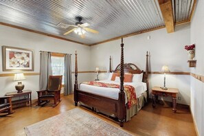 Ein Bett mit Baldachin, ein Holzkopfteil, ein Deckenventilator, ein gerahmtes Blumenbild, ein Schaukelstuhl und ein Nachttisch mit einer Lampe.