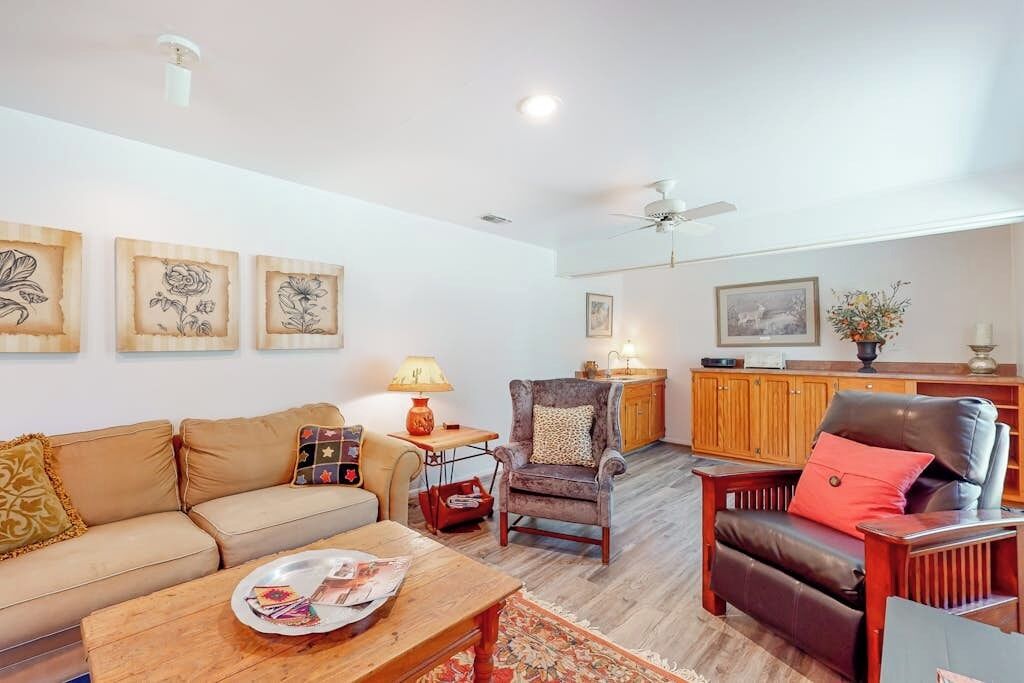 Una sala de estar con sofá, sillón, mesa de centro y una pequeña cocina con un armario y una lámpara.