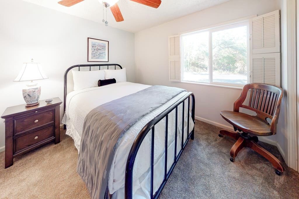 A bedroom with a bed, a nightstand, a chair, a window with shutters, and a ceiling fan.