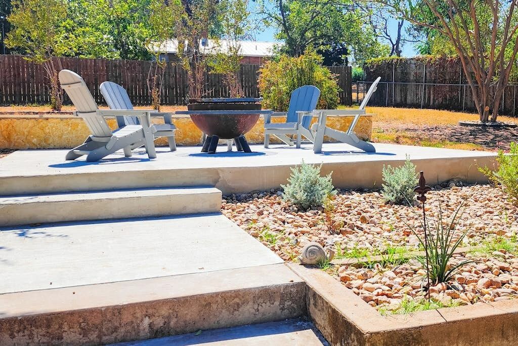 An outdoor patio with a fire pit, three Adirondack chairs, and a concrete step leading to a garden area with rocks and plants.