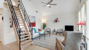 A spacious living room with a wooden staircase, a ceiling fan, a flat-screen TV, and a large window with blinds. - Peaceful, Modern Home Close to Lago Vista With Large Deck & Balcony (Marble Falls)