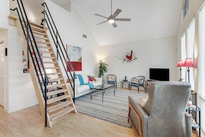 A spacious living room with a wooden staircase, a ceiling fan, a flat-screen TV, and a large window with blinds. - Peaceful, Modern Home Close to Lago Vista With Large Deck & Balcony (Marble Falls)