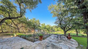 A stone patio with a table and chairs, surrounded by trees and a fenced garden area. - Peaceful, Modern Home Close to Lago Vista With Large Deck & Balcony (Marble Falls)