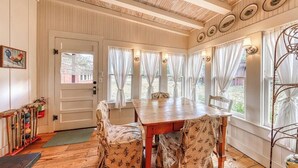 A dining area with a wooden table and patterned chairs, large windows with curtains, and a ceiling with exposed beams. - Historic Farmhouse With Lush Covered Patio - Near Wineries & Main St (Fredericksburg)