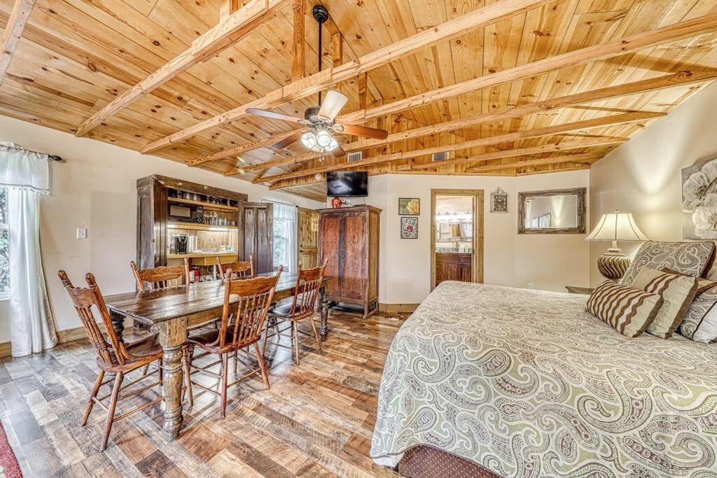 A spacious room with a wooden ceiling, a dining area with a wooden table and chairs, a bed with patterned bedding, and a television mounted on the wall.