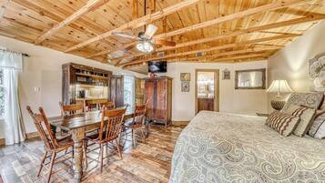 A spacious room with a wooden ceiling, a dining area with a wooden table and chairs, a bed with patterned bedding, and a television mounted on the wall.