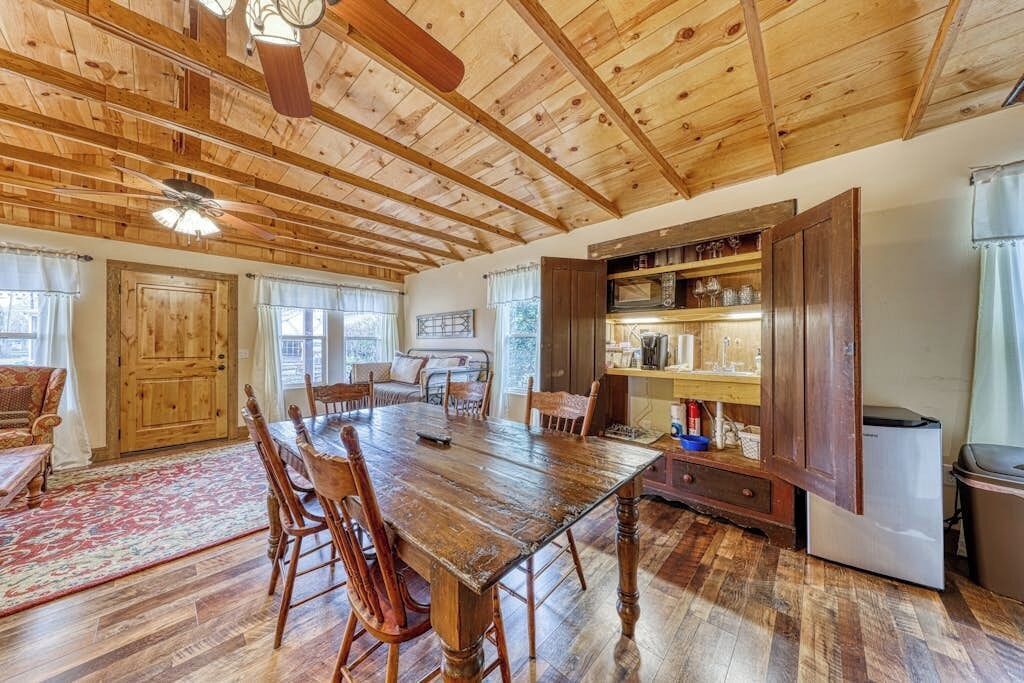 A rustic kitchen with wooden beams, a dining table, chairs, a refrigerator, and a built-in shelf with glassware.