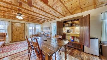 A rustic kitchen with wooden beams, a dining table, chairs, a refrigerator, and a built-in shelf with glassware.
