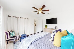 Un dormitorio con cama, ventilador de techo, televisor, sillón y cómoda.