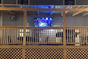 Una terraza de madera con barandilla de celosía, una pantalla de televisión mostrando un programa y un ventilador montado en la pared.