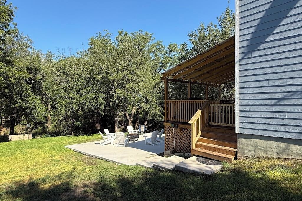 A covered outdoor seating area with a wooden deck, a concrete patio, and a view of a tree-lined backyard.