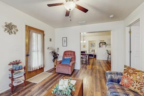 Ein Wohnzimmer mit Deckenventilator, Holzboden, einem rosa Sessel, einem blauen Sofa und einem Essbereich im Hintergrund.
