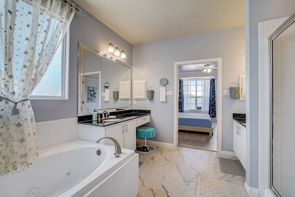 A bathroom with a bathtub, a sink, and a mirror. There is a door leading to a bedroom with a bed and a ceiling fan.