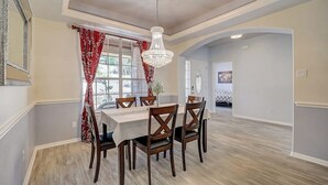 A dining area with a table set for six, wooden chairs, a chandelier, and a view of a garden through a window with red curtains. - Spacious, Dog-friendly Home With Two Decks, Views, Grill, W/D & Community Pool (Leander)