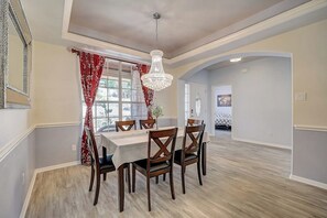 A dining area with a table set for six, wooden chairs, a chandelier, and a view of a garden through a window with red curtains. - Spacious, Dog-friendly Home With Two Decks, Views, Grill, W/D & Community Pool (Leander)