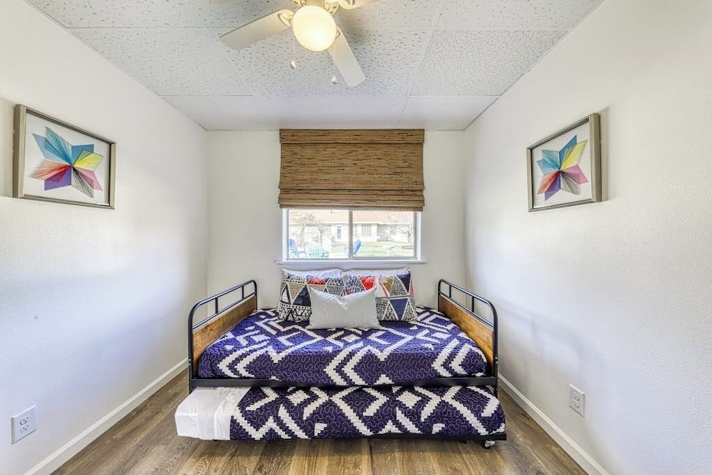 Ein kleines Schlafzimmer mit einem Bett, einem Deckenventilator, einem Fenster mit Jalousien und zwei gerahmten Kunstwerken an den Wänden.