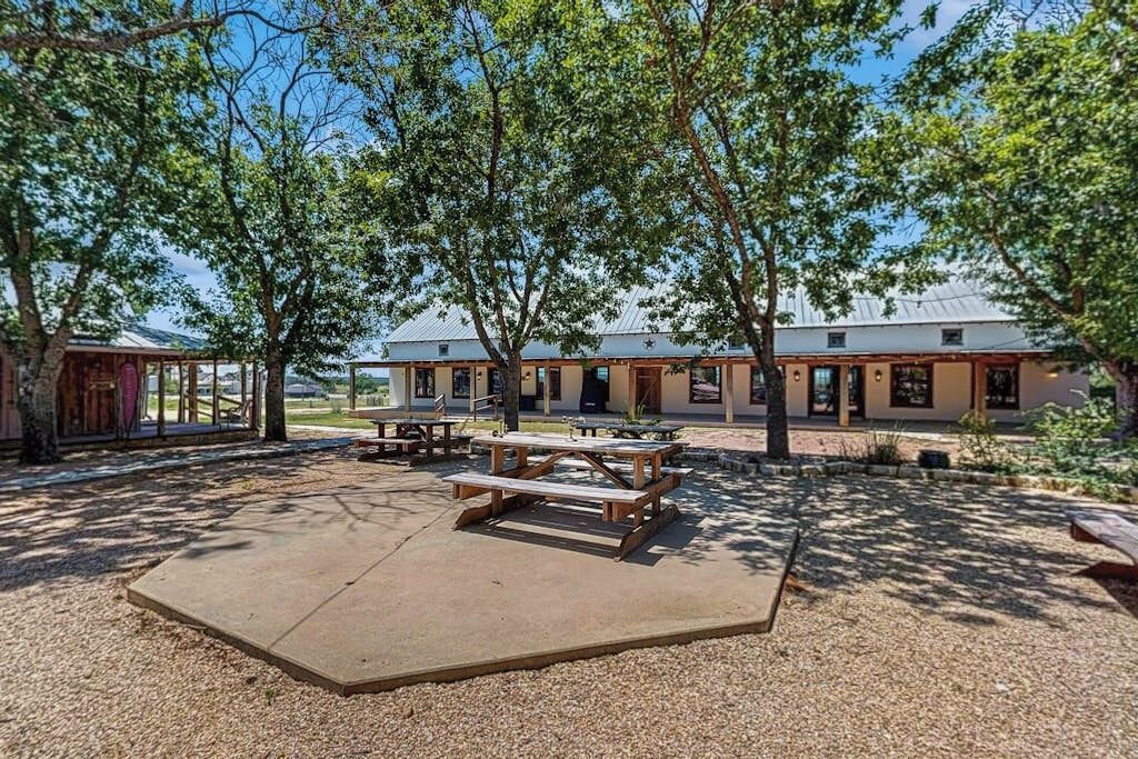 A covered outdoor area with picnic tables, a building with a metal roof, and trees providing shade.