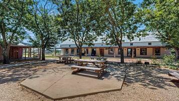 A covered outdoor area with picnic tables, a building with a metal roof, and trees providing shade.