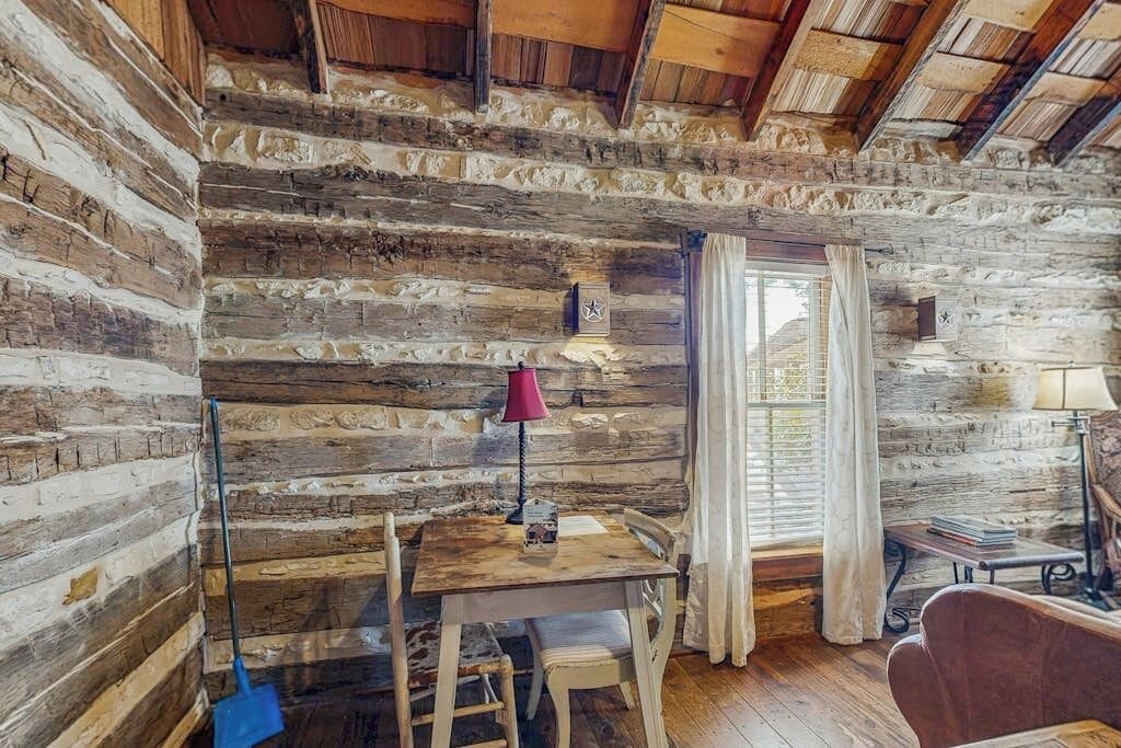 A rustic log cabin interior with a wooden table, chairs, a lamp, and a window with curtains.
