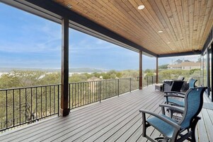 Um deck coberto ao ar livre com piso de madeira e guarda-corpo de metal, com mobiliário para área externa e vista para colinas distantes.