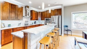 A modern kitchen with wooden cabinets, a marble island, and bar stools. - Bright & Spacious Family Home With Firepit, Lake Views, Furnished Deck, Free Wifi (Leander)