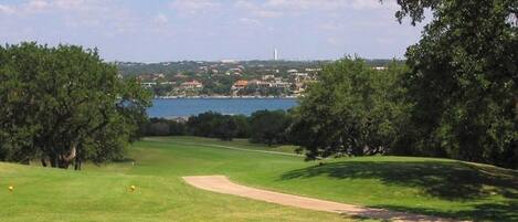 A golf course with a water body in the background, surrounded by trees and buildings.