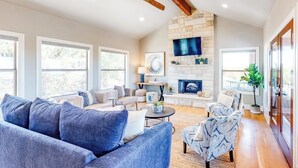 A spacious living room with a stone fireplace, large windows, and a ceiling fan. - Bright & Spacious Family Home With Firepit, Lake Views, Furnished Deck, Free Wifi (Leander)